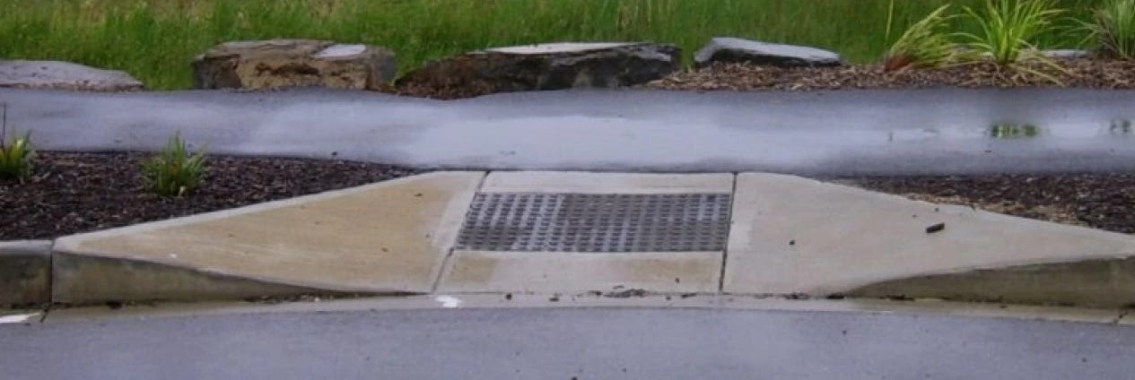 A photo of a sidewalk curb cut designed for wheelchair accessibility. The concrete ramp slopes down from the sidewalk to meet the street, with a textured metal drainage grate in the center for traction and water runoff. The surrounding area includes patches of mulch with small plants and a grassy field in the background. The pavement is wet, indicating recent rain.