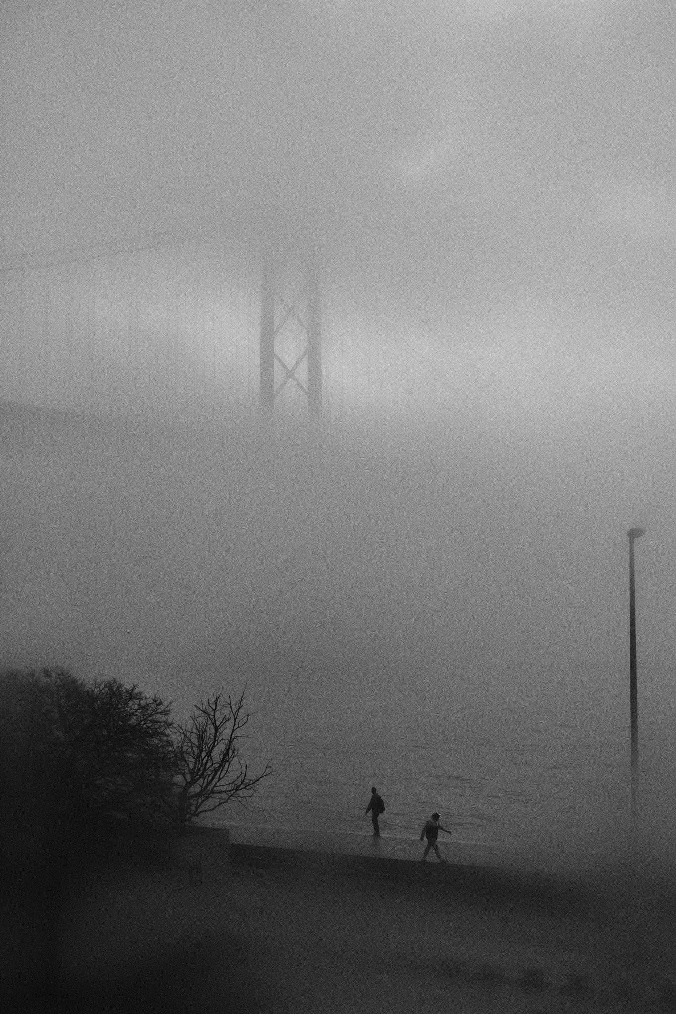 Two people walking by the Tejo river with the 25 de Abril bridge is almost lost in fog in the background.