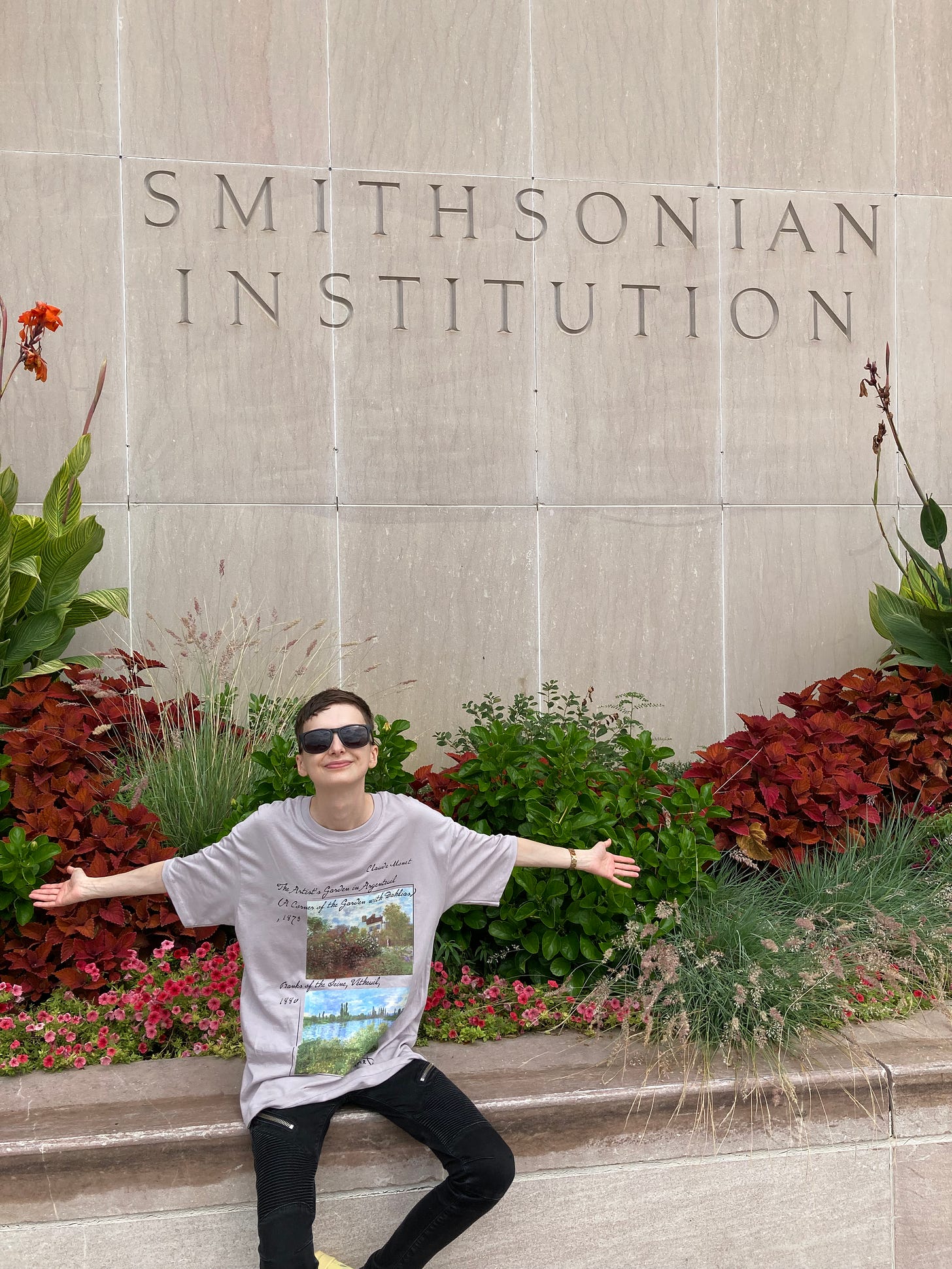 Beck sitting on a planter outside of the museum of American history. The wall is etched with Smithsonian Institution. Beck has their arms spread wide, claiming the space while smiling.