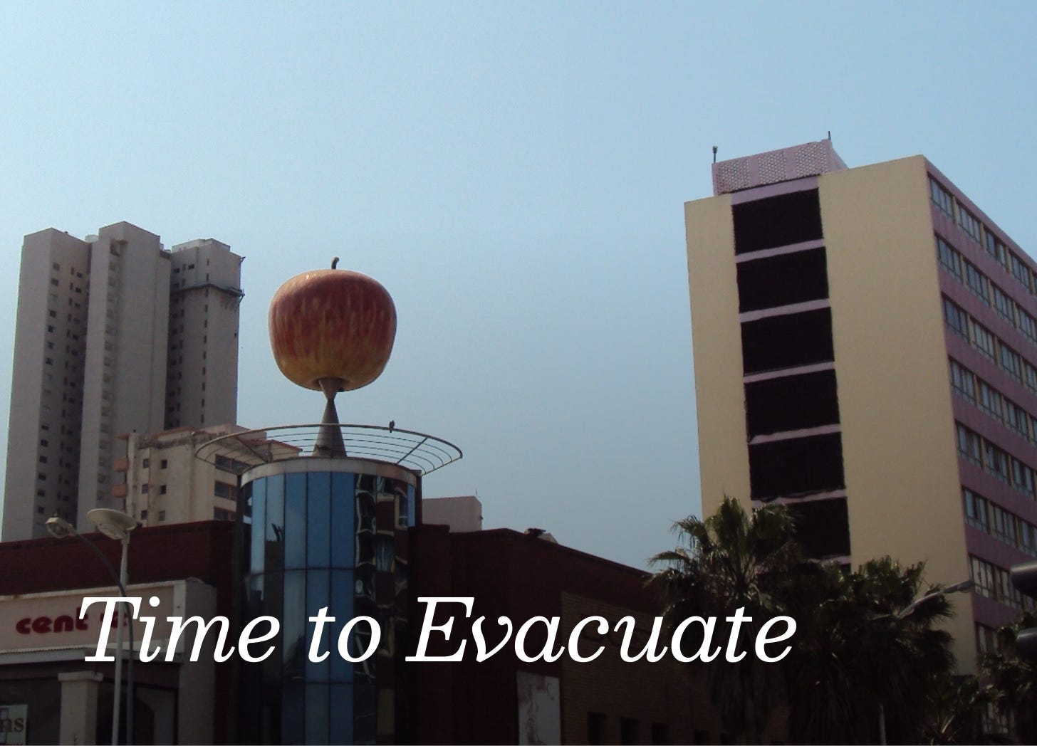 Mid-century modern buildings, with a huge red apple on top of a round building. A light blue sky is behind.