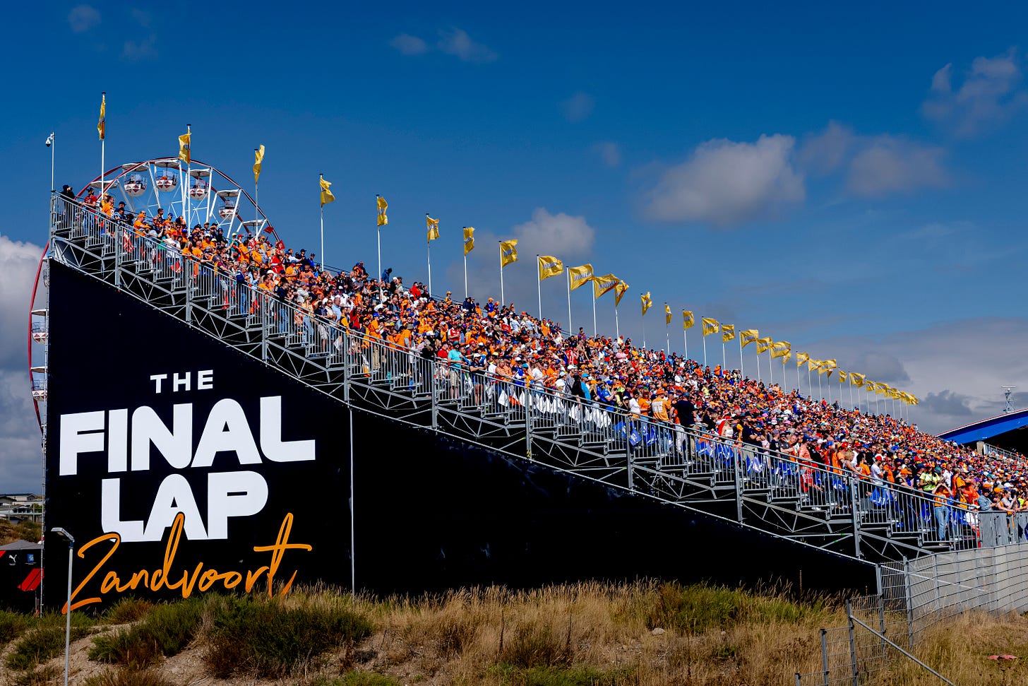 Fans, mostly wearing orange, in a grandstand against a blue sky backdrop with the words 'The Final Lap Zandvoort' written on the side of the structure.