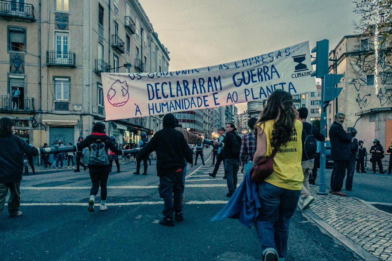 A large banner with the sentence 'governments and companies have declared war on humanity' is unfurled over the praça do Chile during the 23 Nov. protest in Lisbon.