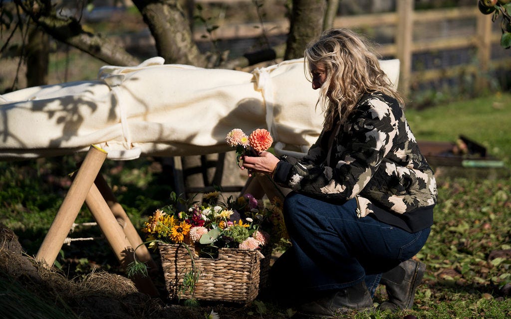 A white woman kneeling next to a basket of flowers. She is holding two flowers and is in front of a white woollen felt shroud.