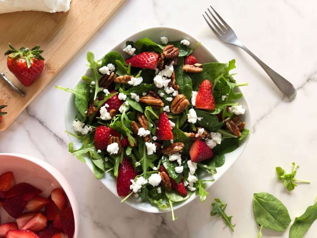 strawberry goat cheese salad in a white bowl with candied pecans and arugula and baby spinach with a bowl of strawberries and goal cheese log in the background