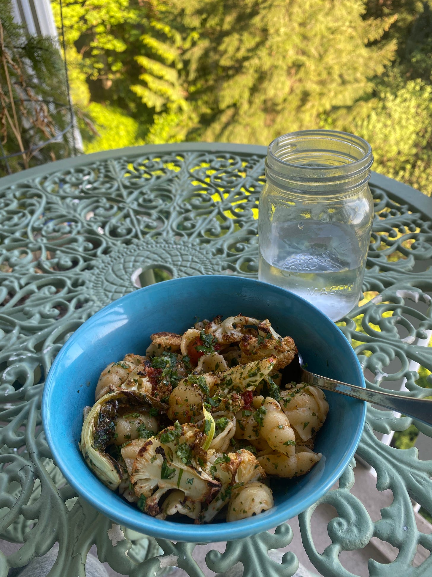 The pasta dish described above, with browned cauliflower florets and leaves, dusted with herbs and drizzled with tomato brown butter. The bowl is sitting on a green metal café table on a balcony, with a sunny, green background below.