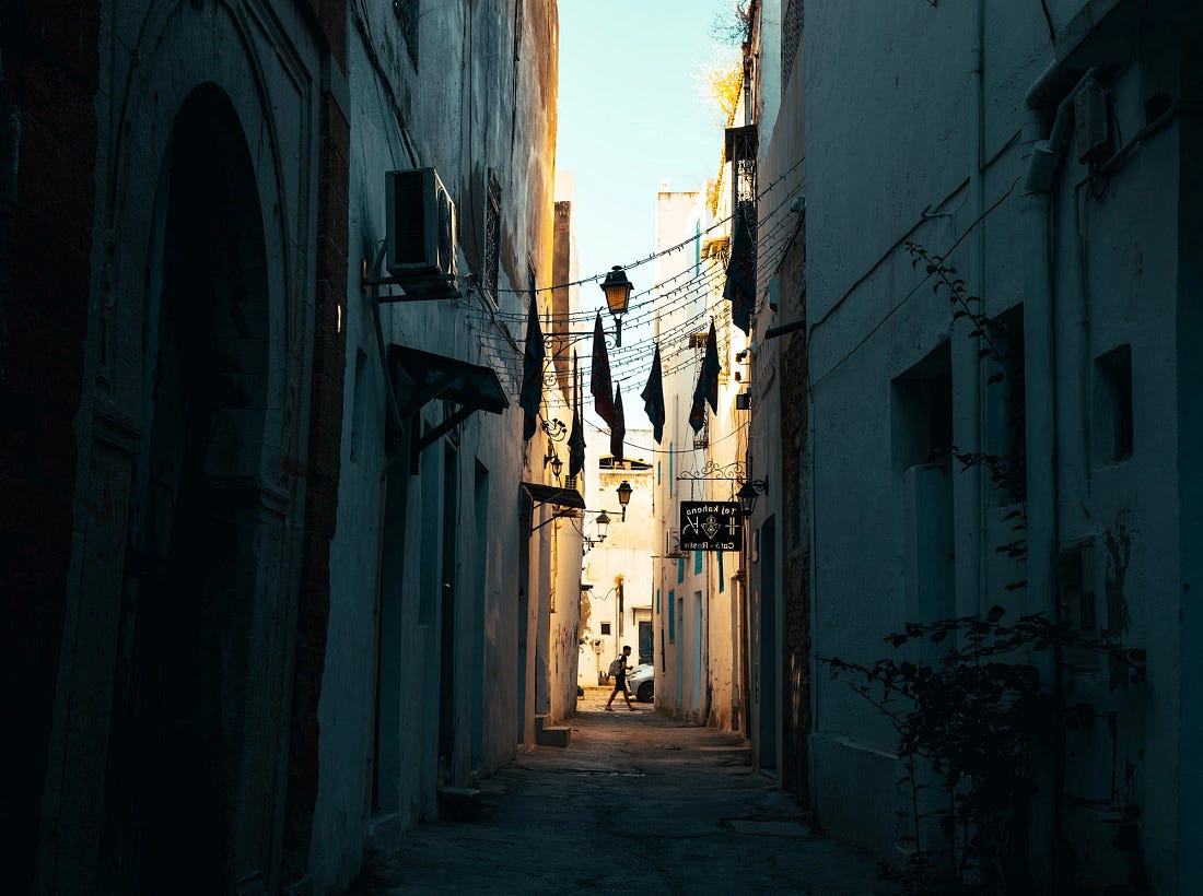 Narrow blue-toned alley in the Tunis Medina at dusk, with whitewashed walls, hanging flags and lights, and a lone pedestrian crossing a sunlit opening at the far end.