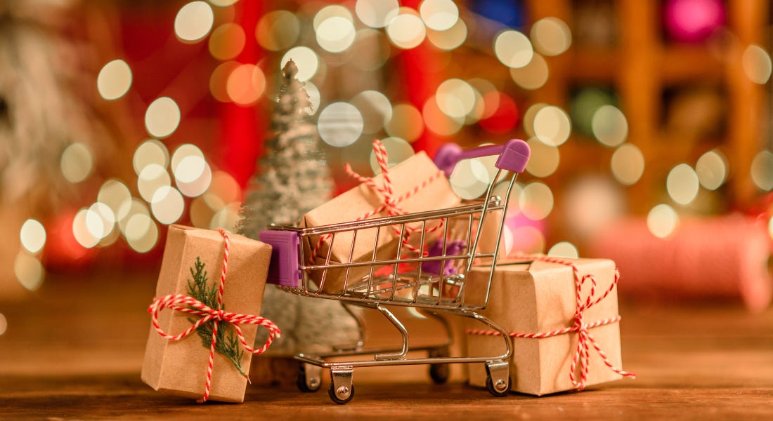 A close-up of a small metal trolley with wrapped presents in it and around it on a table, with lights and a Christmas tree in the background