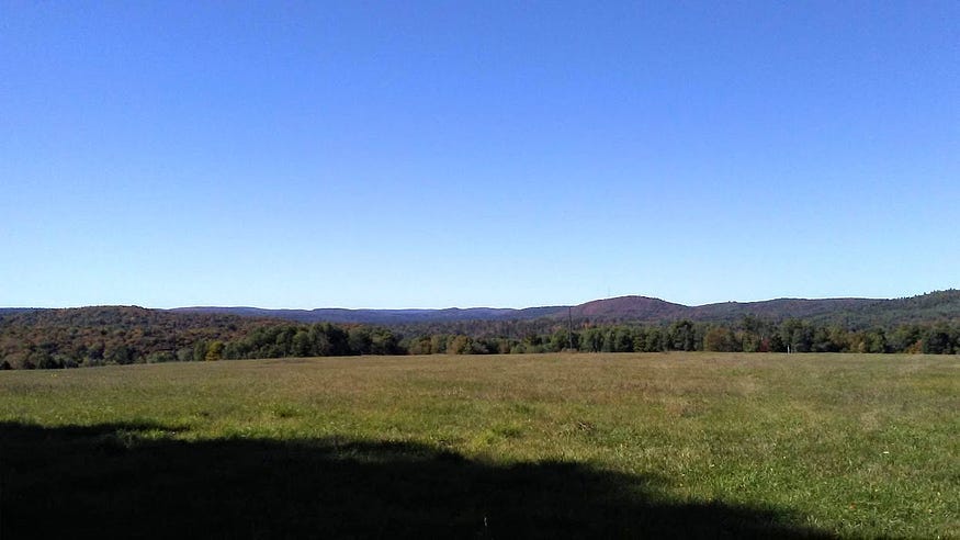 A line of distant hills look purple in the afternoon sun against the blue of the sky. In the foreground a field drops away into a wooded valley.