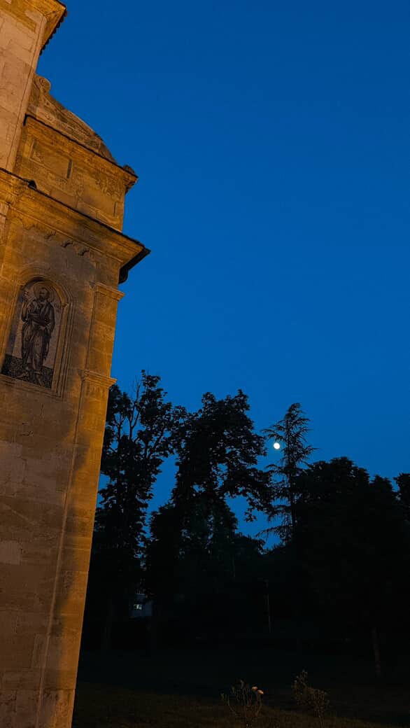 1) A castle tower, to the left, sits in front of a small forest at blue hour on a full moon. 2) A castle-like building featuring spires & ornate window decorations in sandstone & salmon colours. 3) At night, a lit red candle on a desk overlooking a cityscape through a window. 4) A stone pathway to a castle, lined with large trees, brush, and white flowers.