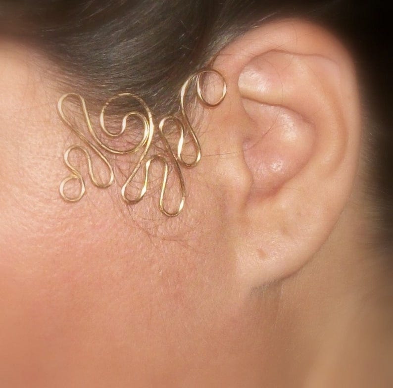 May include: Gold ear cuff with a delicate, intricate design. The cuff is made of thin wire and has a flowing, organic shape.