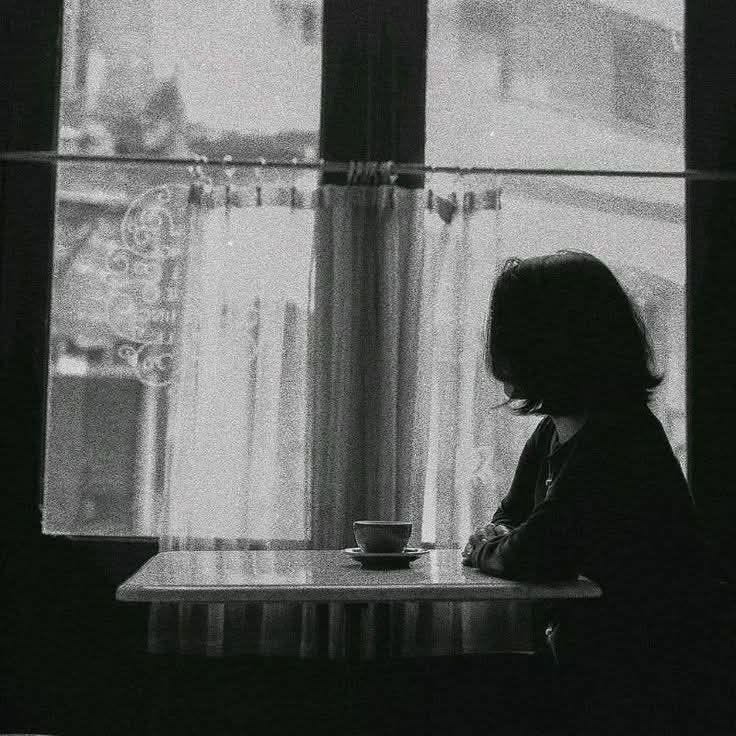 This may contain: a woman sitting at a table in front of a window with a cup on it