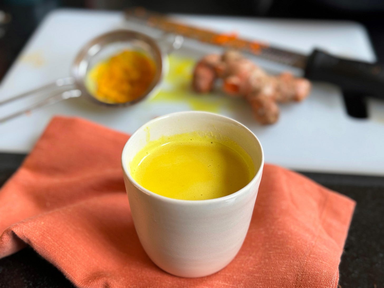 A cup of golden milk (haldi doodh) in a white ceramic cup on a coral-colored napkin, with fresh turmeric root, a Microplane grater and a strainer on a cutting board in the background