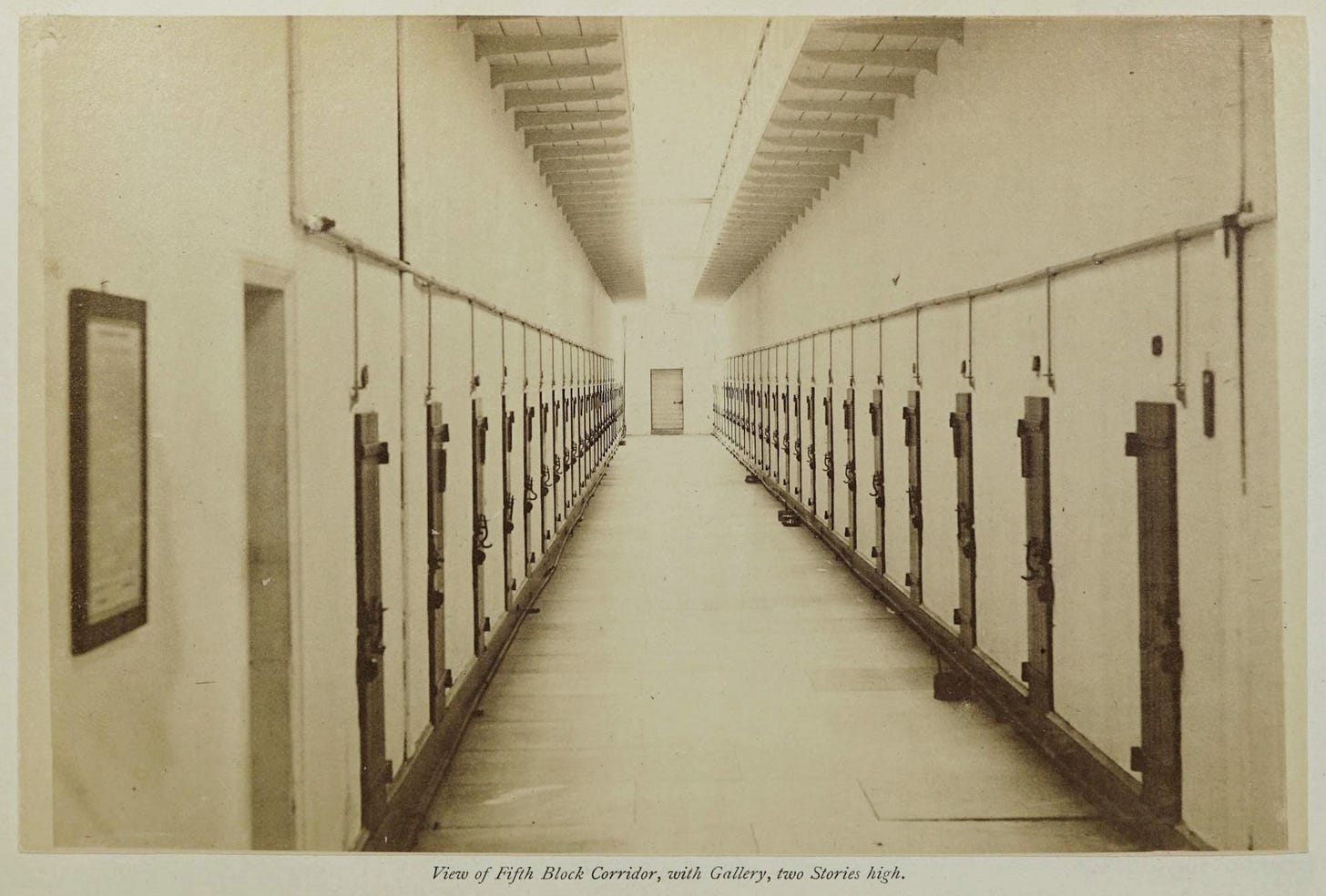A black and white public domain image of a long corridor inside a prison with cell doors lining both sides of the walls and a single door at the far end of the hallway. A black and white public domain image of a long corridor inside a prison with cell doors lining both sides of the walls and a single door at the far end of the hallway.