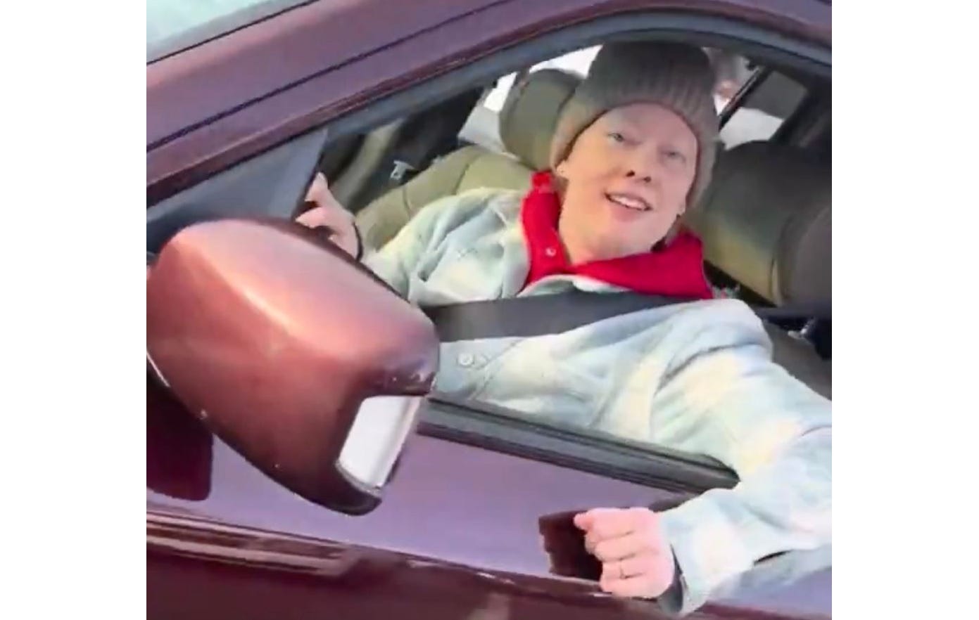 video screenshot of Renee Good in the driver's seat of her maroon Honda Pilot SUV. She wears a tan knit cap, and a light grey plaid winter coat over a red hoodie. She smiles as she speaks to the ICE agent who's recording her from his cell phone. 