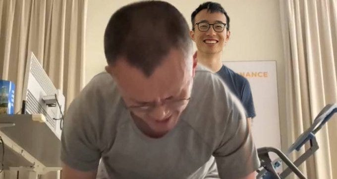 Two men in a room, one performing a push-up in a gray shirt, the other standing and smiling in a blue shirt. An exercise bike and a whiteboard with "Binance" are visible in the background.