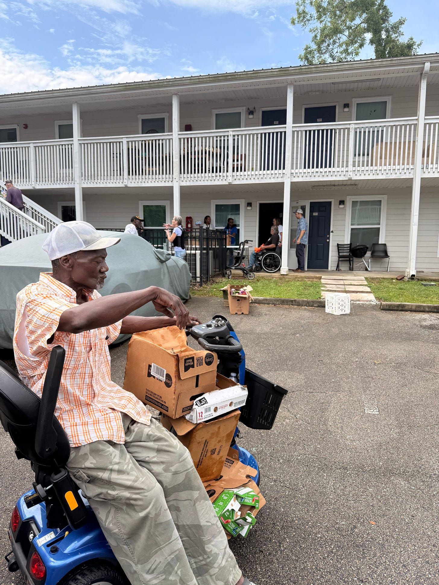 In a parking lot outside a two-story motel-style building with white railings and blue doors, a man in a white cap and orange plaid shirt sits on a blue mobility scooter loaded with cardboard boxes. Several people stand and talk near a ground-floor doorway, with a person in a wheelchair nearby, under a partly cloudy sky. In a parking lot outside a two-story motel-style building with white railings and blue doors, a man in a white cap and orange plaid shirt sits on a blue mobility scooter loaded with cardboard boxes. Several people stand and talk near a ground-floor doorway, with a person in a wheelchair nearby, under a partly cloudy sky.