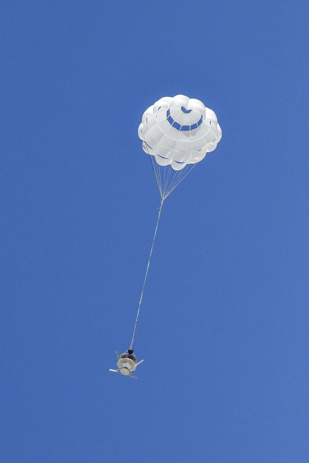 A parachute carrying a capsule is fully open against a canvas of deep blue sky. A parachute carrying a capsule is fully open against a canvas of deep blue sky.