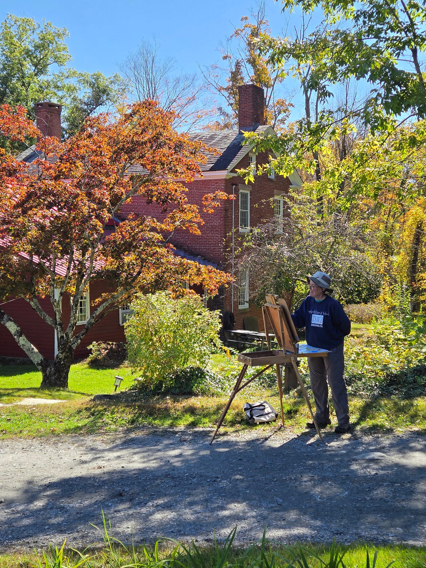  A person in a hat and dark clothing paints on an easel outdoors, positioned on a gravel path with a red brick building and trees with autumn foliage in the background.