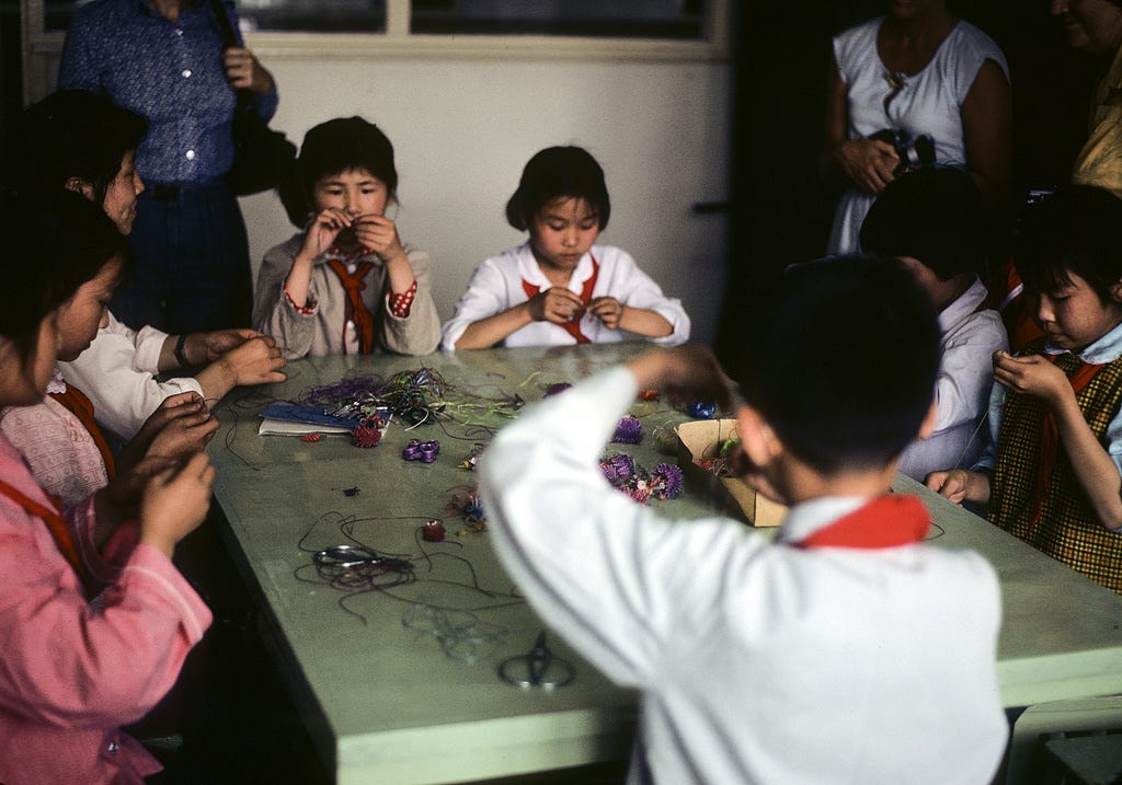 1979 color photograph of several Chinese children intently making beaded crafts around a light green table. On the table are colorful string and  beads, scissors, and other supplies. The kids wear a range of outfits, but all have matching red bandana ties. A few adults stand against the wall. Only their torsos are pictured.