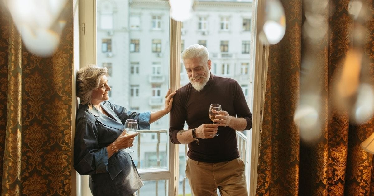 A senior couple enjoys wine and laughter by balcony windows inside a cozy apartment A senior couple enjoys wine and laughter by balcony windows inside a cozy apartment
