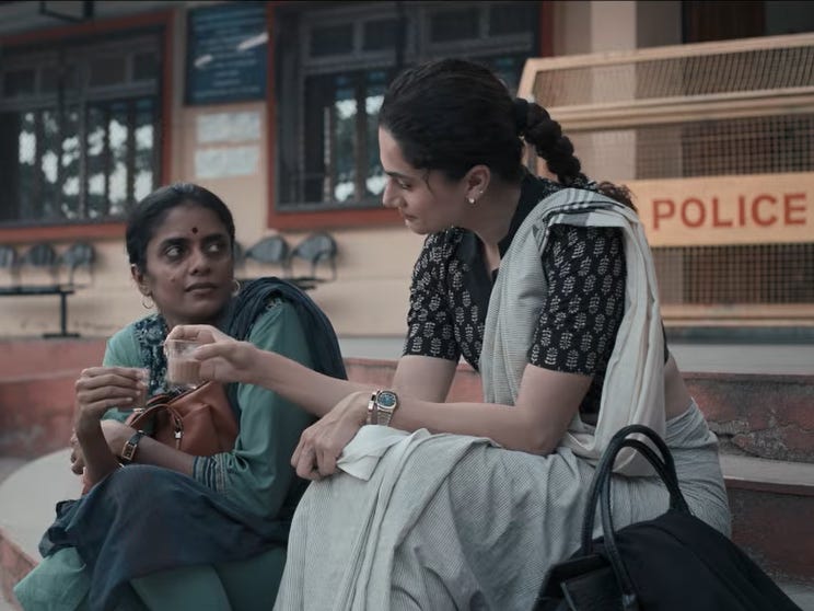 A still from Hindi film, ‘Assi (2026)’. A still from Hindi film, ‘Assi (2026)’. Parima (Kani Kusruti) sits on the left on the steps outside a building which might be a court house or a police station. She is wearing a teal kurta with a dark dupatta, a bindi, and hoops, clutching a brown handbag as she looks towards Raavi (Taapsee Pannu). Raavi is sitting on a step or two above Parima. She is wearing in a light grey sari with a black patterned blouse, her hair pulled into a long braid, wearing small earrings and a wristwatch, leaning in and looking at Parima with quiet concern. A black bag lies on her side. They both have small tea glasses in their right hands. A red “POLICE” sign is visible on a yellow barricade behind them.