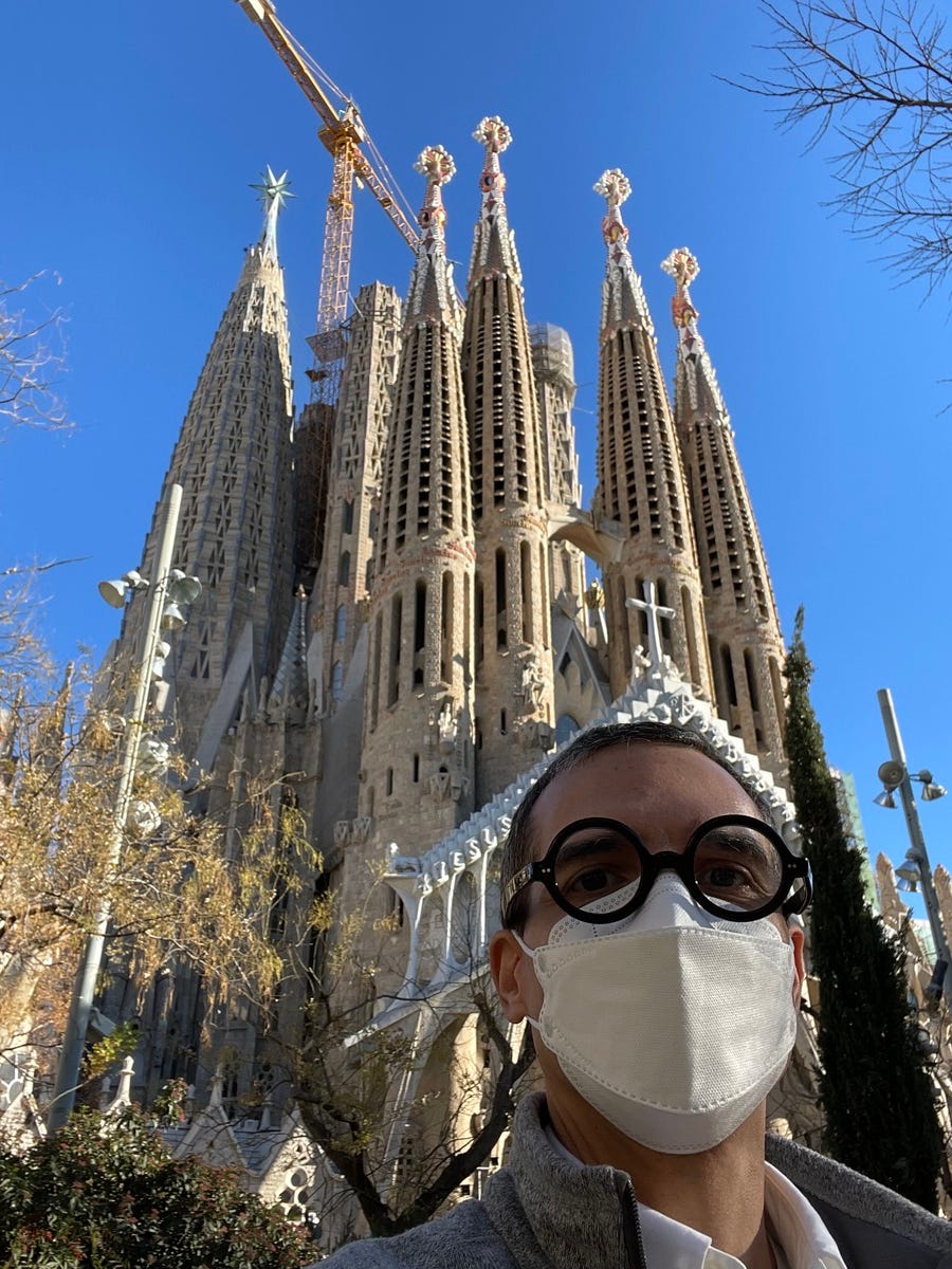 A person wearing glasses and a face mask stands in front of the Sagrada Familia in Barcelona, with its distinct spires and a construction crane visible. It’s about time I shared a selfie in the course.