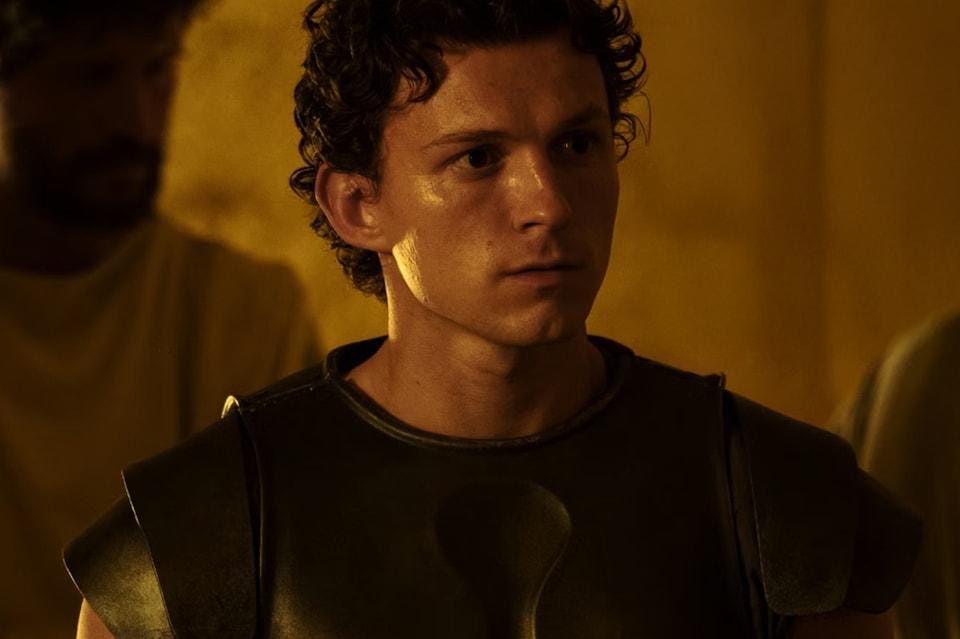 Telemachus, played by Tom Holland, wears dark armor and looks tense in warm interior light. Telemachus, played by Tom Holland, wears dark armor and looks tense in warm interior light.