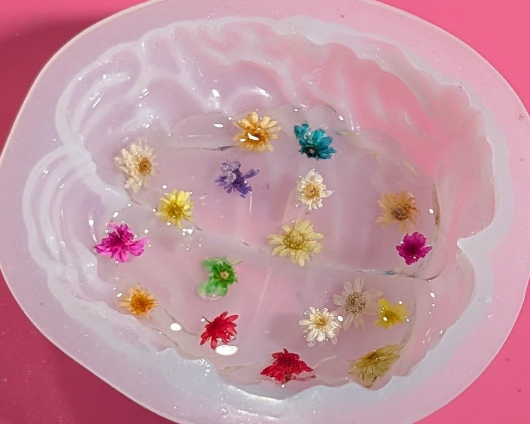 A top-down view of a clear silicone mold shaped like a brain, resting on a bright pink surface. The mold is filled with a clear resin and multiple tiny, colorful dried flowers—yellow, pink, teal, purple, red, and white—are suspended and preserved inside, looking like thoughts being planted.
