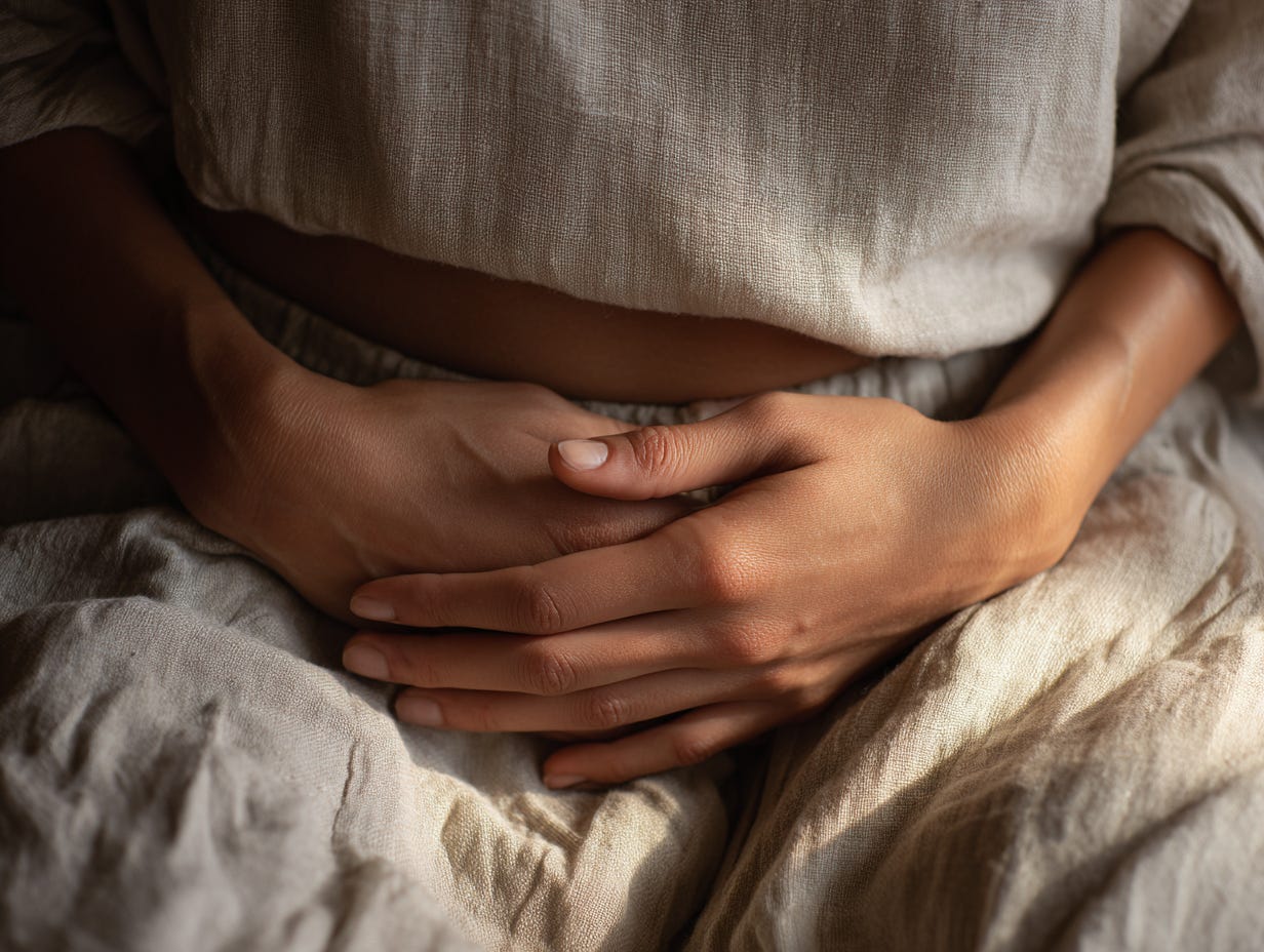 Close-up of hands resting over the lower belly on soft linen, warm window light creating a calm, grounded mood