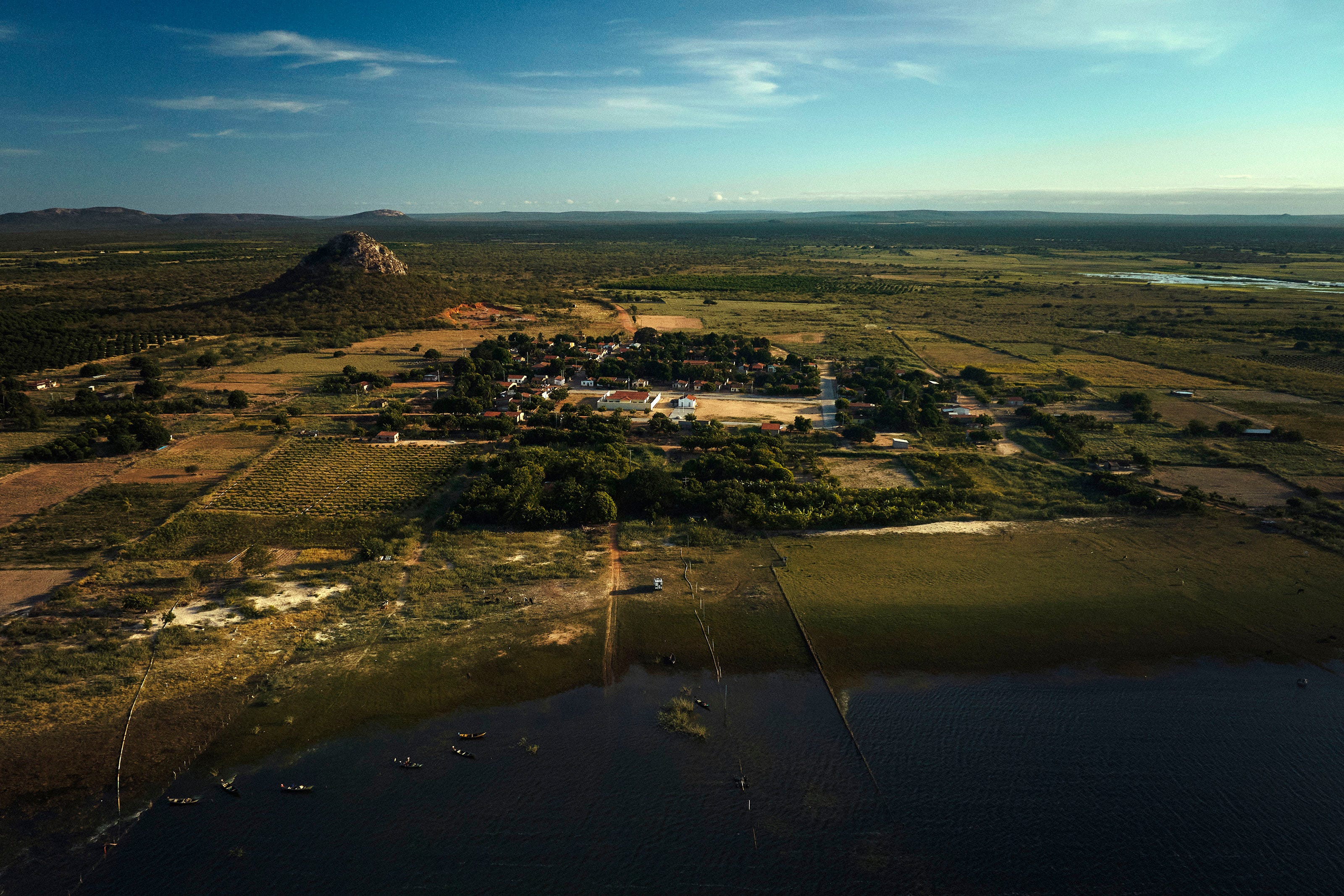 Aerial view of a small village surrounded by fields and greenery, with a body of water in the foreground and a distinctive hill rising in the background under a clear blue sky.