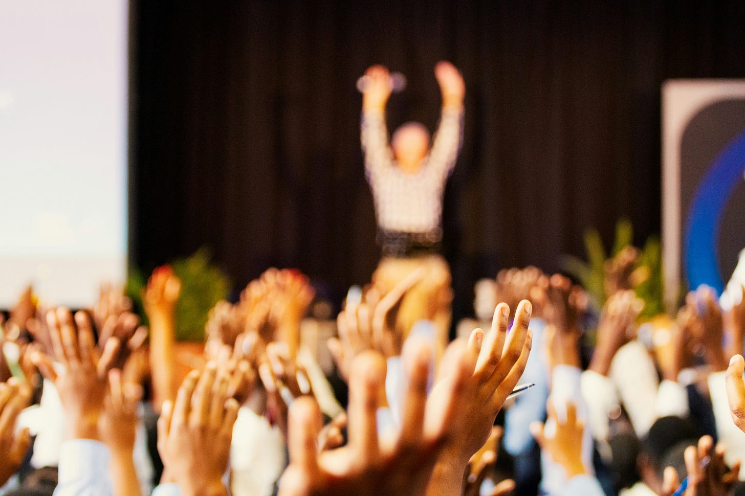 Blurry photo of conference attendees raising hands. Speaker is on stage also raising both hands towards the ceiling.