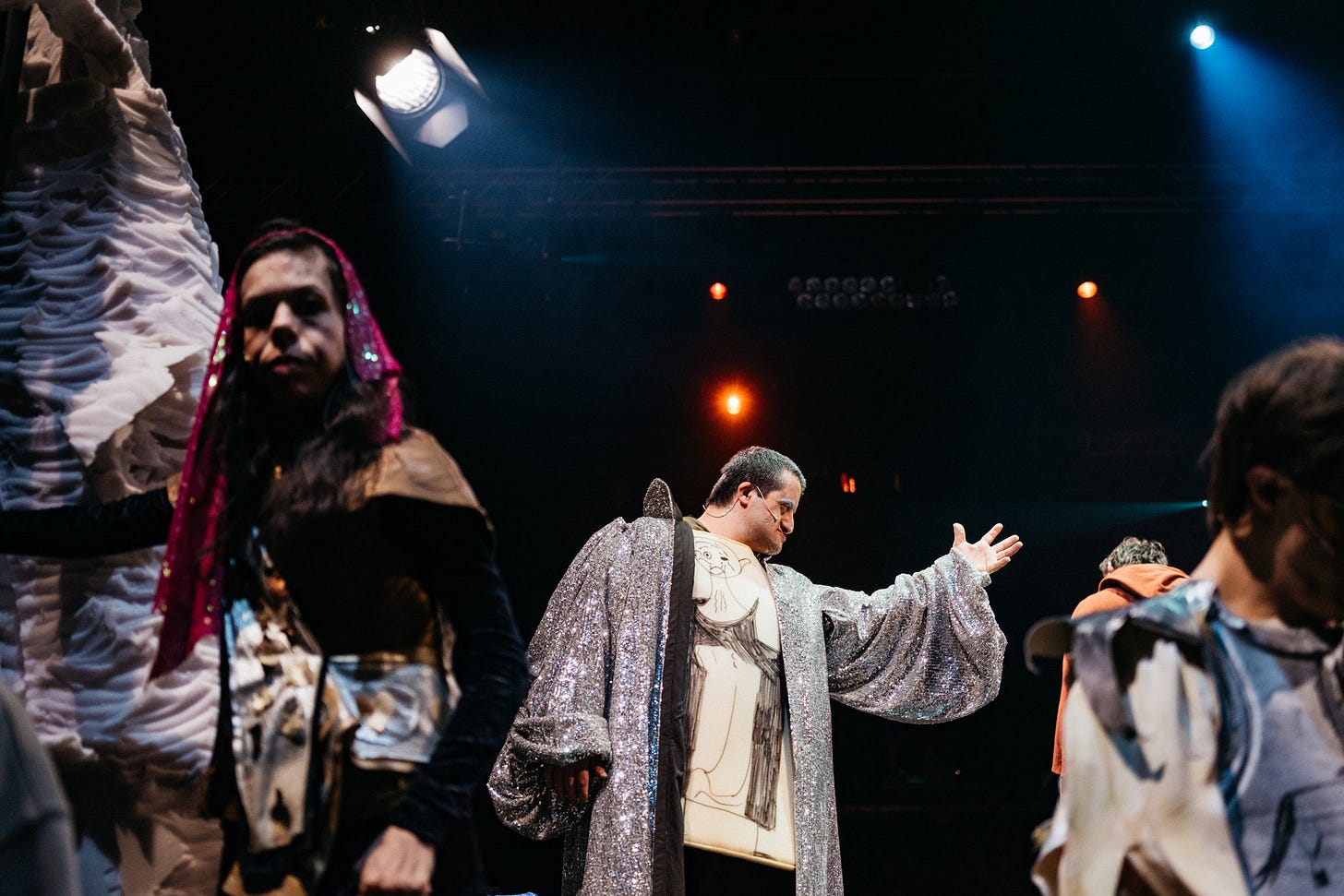 An actor in a silver robe raises his hand in a dramatic gesture