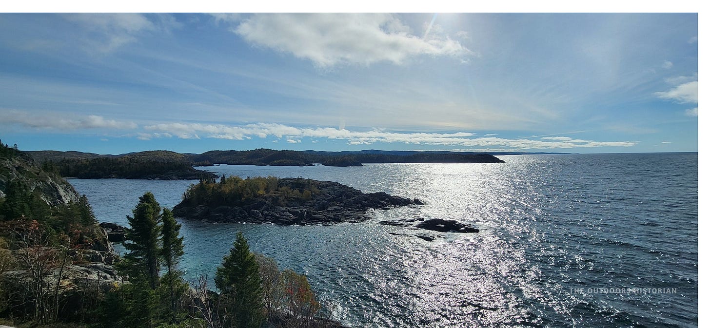 A breathtaking view from a high vantage point overlooking Lake Superior. Rugged, tree-covered islands and rocky points stretch out into the vast, blue water under a bright sky, showing the immense scale of the landscape.