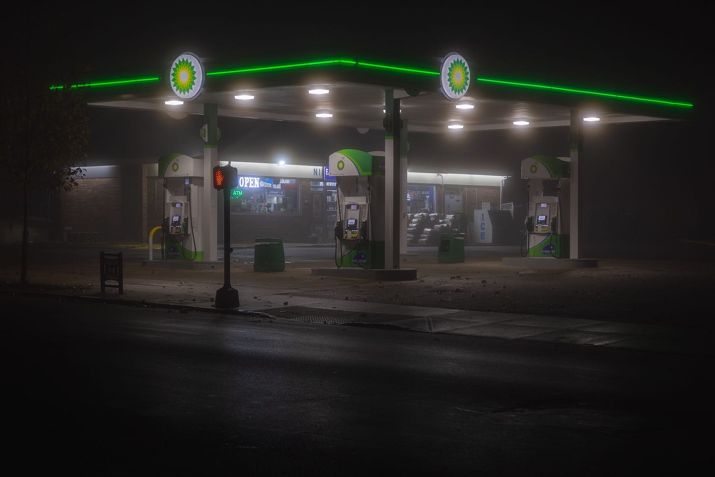 A foggy night at a BP gas station, illuminated by green and white lights with a glowing “OPEN” sign visible in the store window.