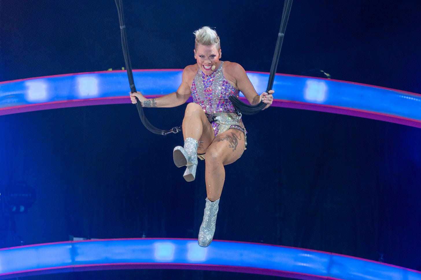 P!nk is seen performing an aerial stunt several feet above the stage. She kicks one foot out while holding onto her harness. She is wearing a sparkly bodysuit and grinning.