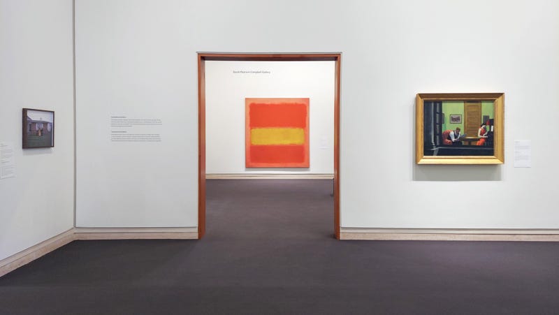 A white walled art gallery with an orange and yellow color field painting in the center, and a painting of two lonely people in New York apartment on the right