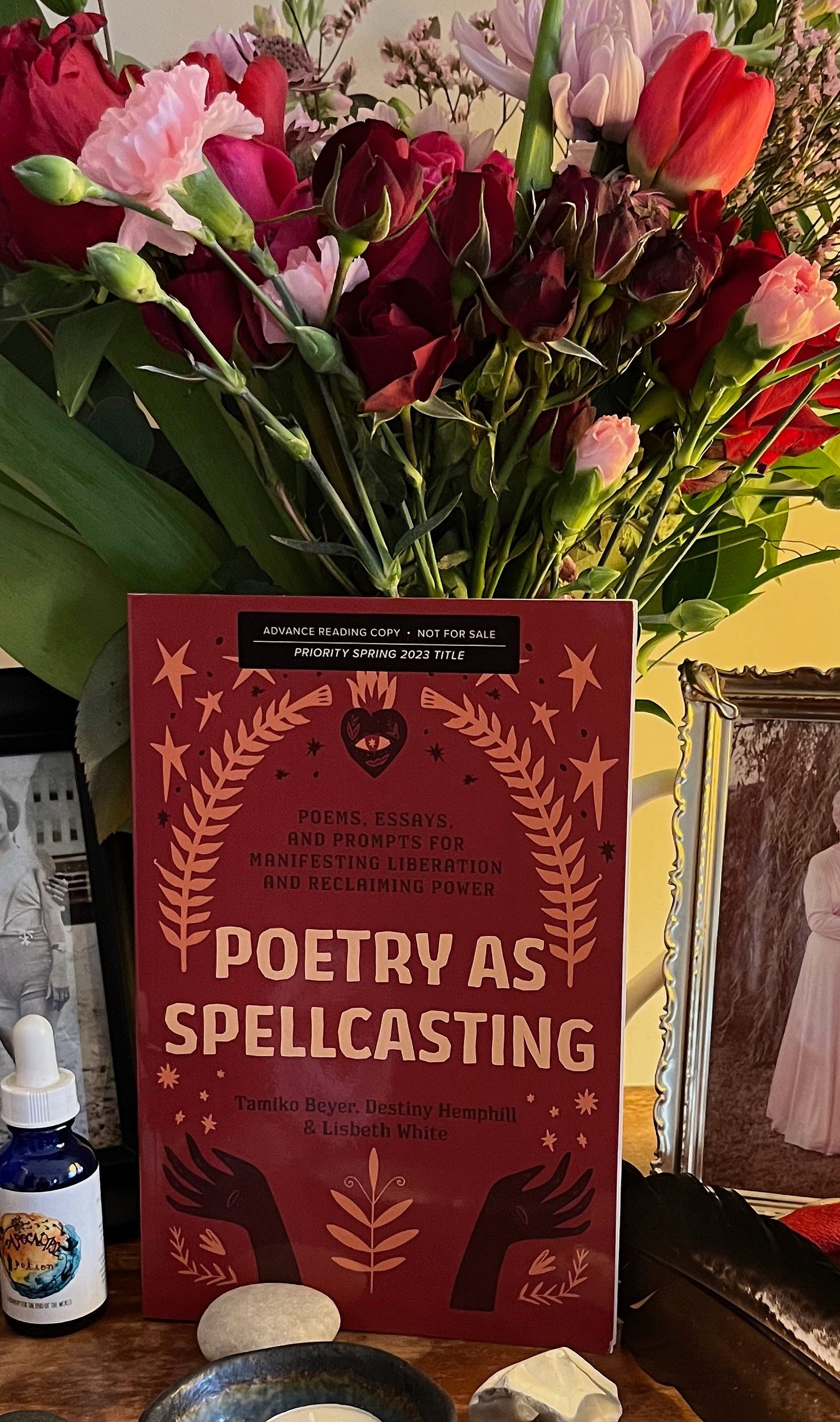 An advanced reading copy of Poetry As Spellcasting is placed on an altar in front of a bouquet of roses, tulips, chrysanthemums, and carnations. The reds and oranges of the books match the colors of the flowers. An advanced reading copy of Poetry As Spellcasting is placed on an altar in front of a bouquet of roses, tulips, chrysanthemums, and carnations. The reds and oranges of the books match the colors of the flowers.