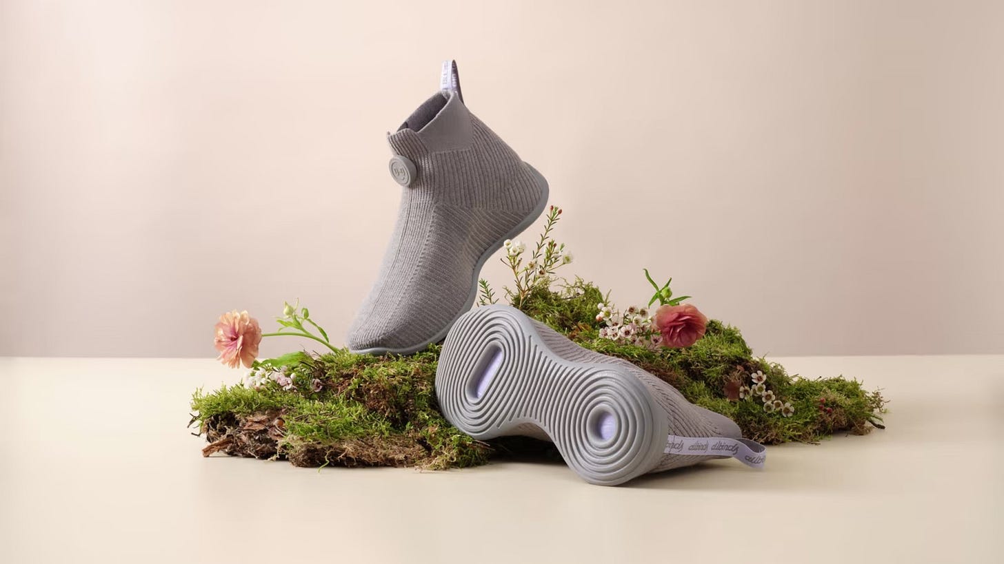 Grey Allbirds moonshot shoes mounted on a cut section of grass with pink flowers against a beige background. Press image.