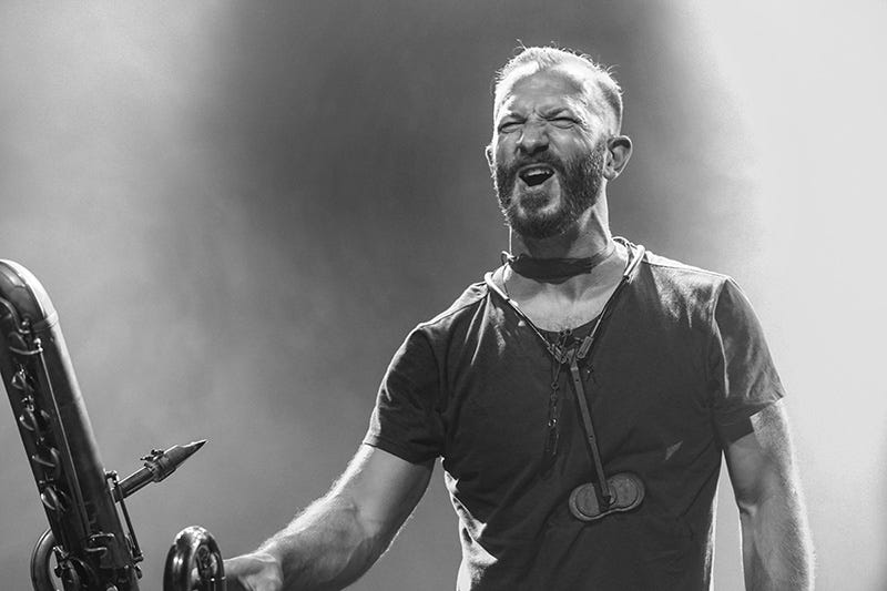 Black-and-white photograph of multi-instrumentalist Colin Stetson smiling with eyes slitted, holding his bass saxophone on a concert stage during a performance Black-and-white photograph of multi-instrumentalist Colin Stetson smiling with eyes slitted, holding his bass saxophone on a concert stage during a performance