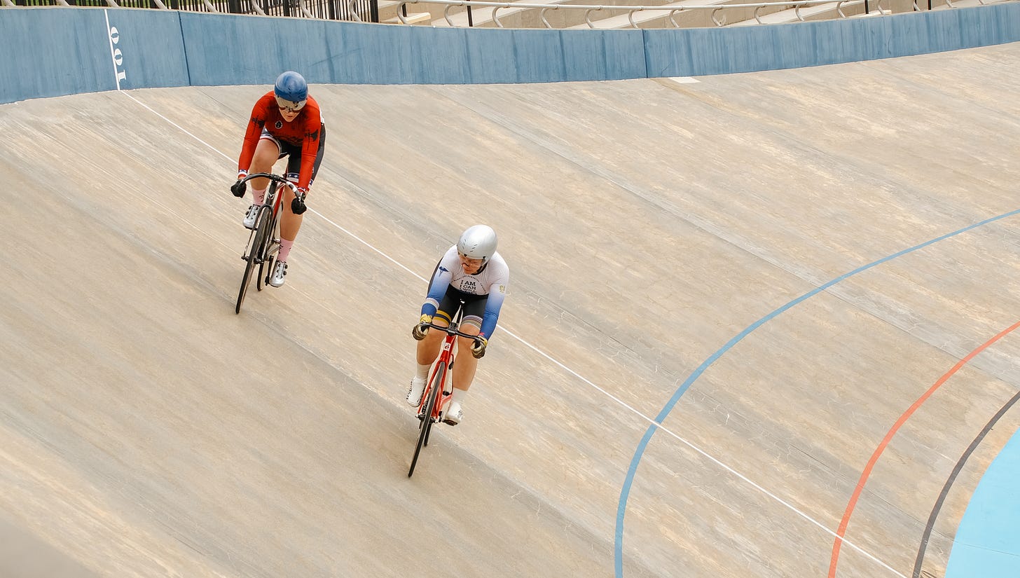 Two track cyclists on the banking of a wooden velodrome, one rider tucked closely behind the other's rear wheel in the draft.