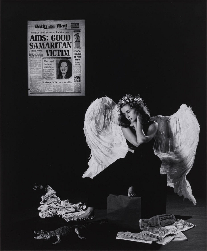 1) A woman with large white angel wings and a floral crown sits pensively amid scattered newspapers and a small faux alligator. A newspaper headline about an AIDS victim is featured on the wall behind the angel. The scene conveys a somber, reflective tone. The angel is holding her head up with her hands. The photo is in black and white, and the angel is barefoot, sitting on a box. 2) A woman wearing large, white angel wings, and a flower crown, sits barefoot pensively on a box. There's a newspaper with David Wojnarowicz on the wall behind her and a small faux alligator on the ground in front of her along with several AIDS and safe sex pamphlets, creating a surreal scene. The photo is in black and white.