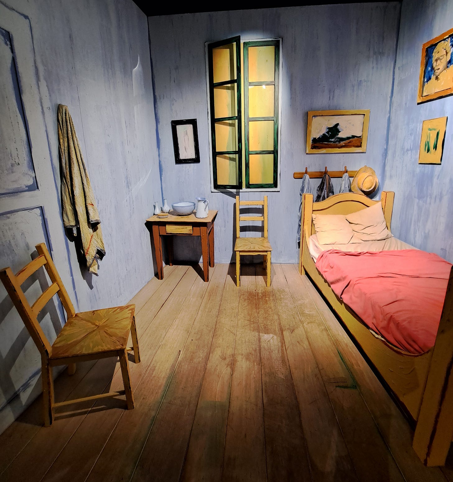 Construction of Van Gogh's painting "The Bedroom at Arles" as seen at Van Gogh: The Immersive Experience. This full-sized replica of the bedroom features the bed on the right, with paintings hung nearby and a row of hooks behind, shuttered windows, a small table and wash basin, two chairs, and a hook on which hangs a painter's cloth.
