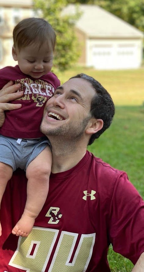 Rikleen carrying a very cute toddler in shorts on his shoulder in a yard