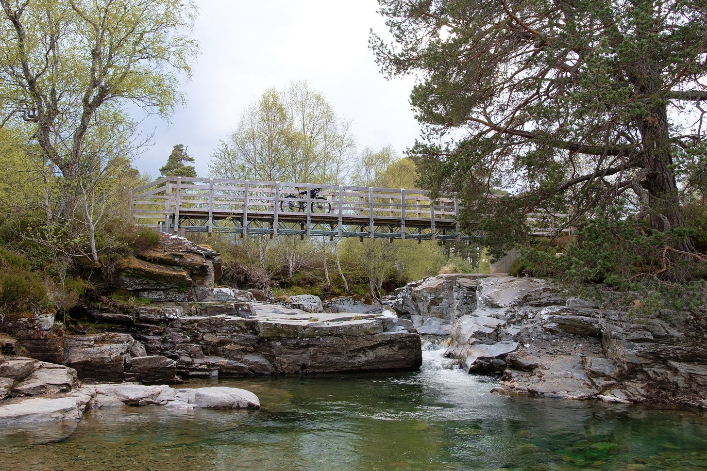 A photo of the wild pools I searched for, with clear green water running through smooth rock shaped by the river. A wooden footbridge crosses above the falls, and a bicycle is nestled beside it. The setting feels secluded, vibrant, and full of life.