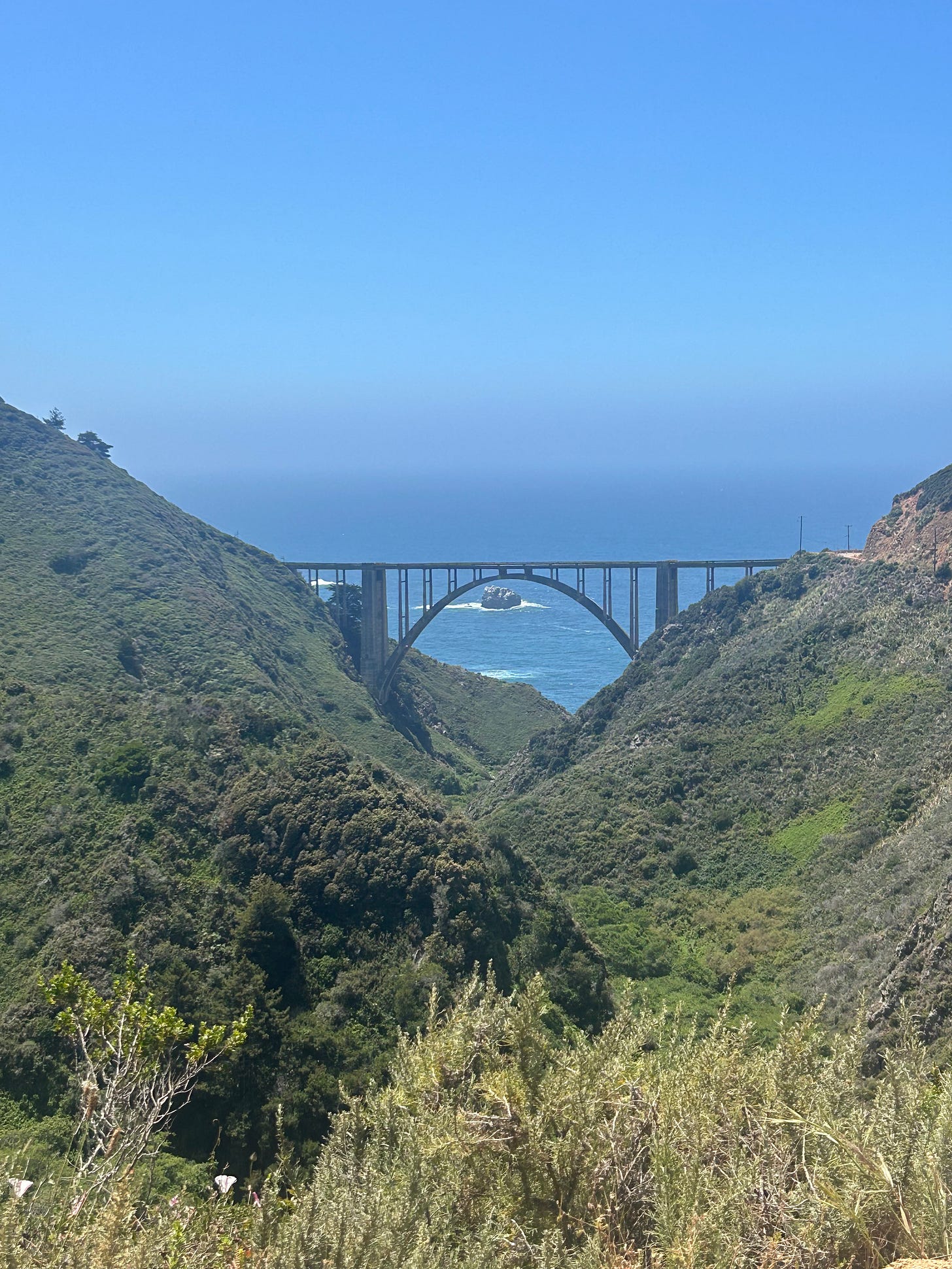 The Bixby Bridge on Highway 1 in Big Sur peeks out from between two mountains. A rock in the ocean sits behind it.