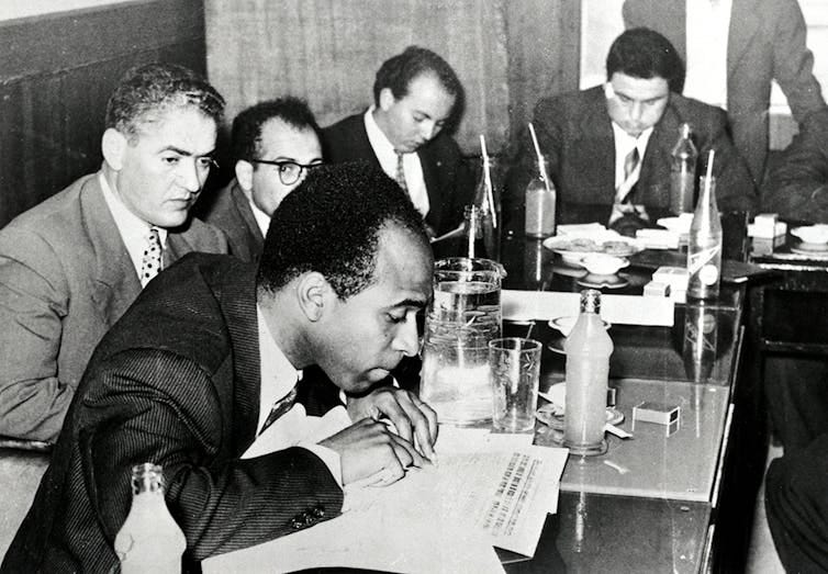 A group of writers, including Fanon, sit around a conference table. A group of writers, including Fanon, sit around a conference table.