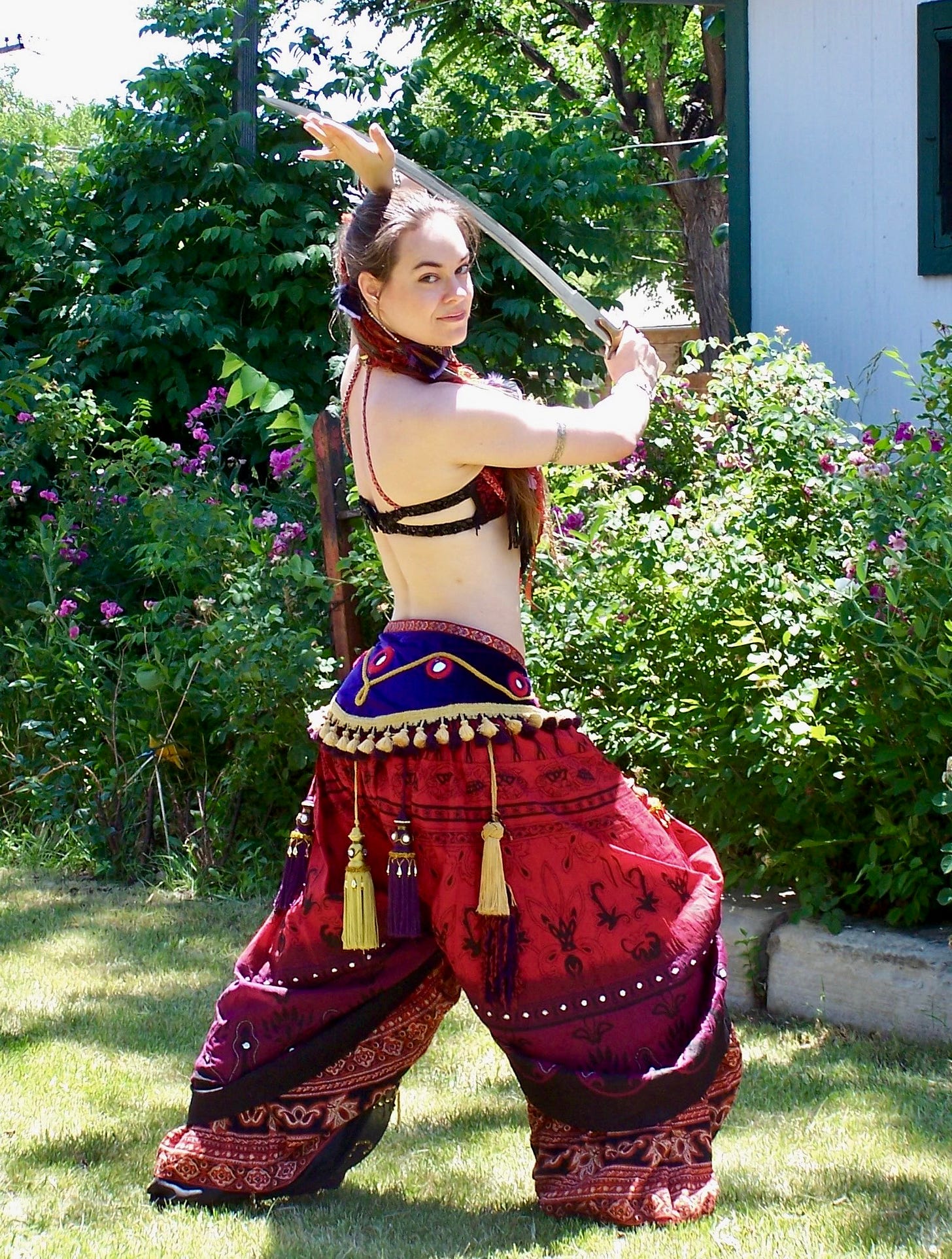 The author in a crimson and purple costume of pants, tassel belt and fringed top. Sword braced to swing, she stares you down over her shoulder with a smile that holds many secrets.