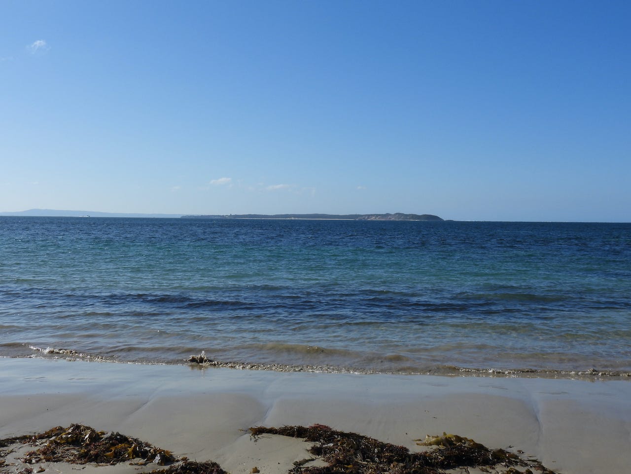 Seascape: an expanse of blue water beyond a sandy beach. Across the water, a headland. (View across the Heads from Point Lonsdale to Point Nepean.