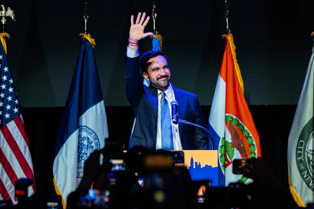 Zohran Mamdani smiles and waves while standing at a lectern onstage.