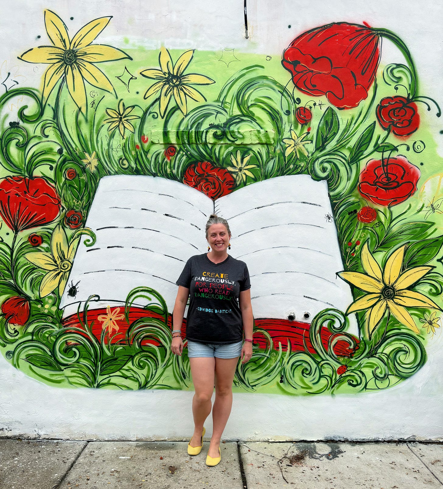 I’m standing in front of an outdoor mural in which a book lies open in a garden of flowers and swirly grass. I’m wearing a t-shirt that says, “Create dangerously, for people who read dangerously. -Edwidge Danticat”